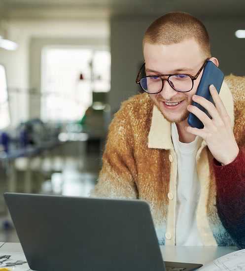 A man in glasses and a cozy sweater talks on a smartphone while working on a laptop in a modern workspace, smiling as he looks at the screen.